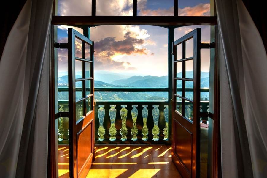 Hotel balcony with open doors and a sunset view over green hills and distant mountains